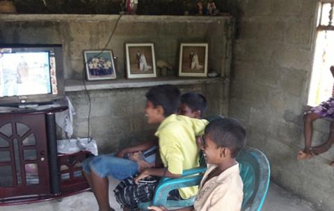 The children are watching in front of their television. 3 are sitting on plastic chairs and 2 are sitting on the window sill.