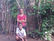 A boy kneeling on the ground and behind him is a girl standing in front of a stick fence.