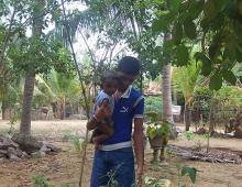 A teenage boy holding his younger baby sibling with one arm. He is surrounded by shade of tall green plants and trees.