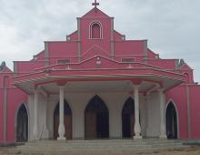 A pink church with 4 white pillars on the porch. At the very top of the church is a cross.